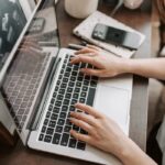 From above of unrecognizable woman sitting at table and typing on keyboard of computer during remote work in modern workspace